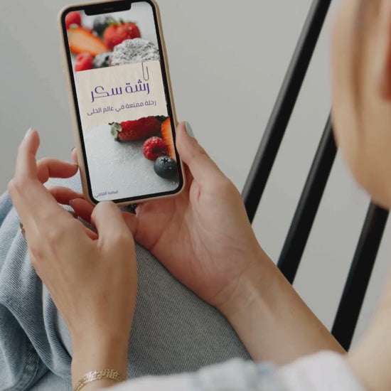 امرأة تمسك هاتفًا يعرض غلاف كتاب إلكتروني بعنوان 'رشة سكر | دليلك للحلويات الشرقية والغربية تعلم اجمل الوصفات' مع صورة لحلويات بالتوت والسكر البودرة.
