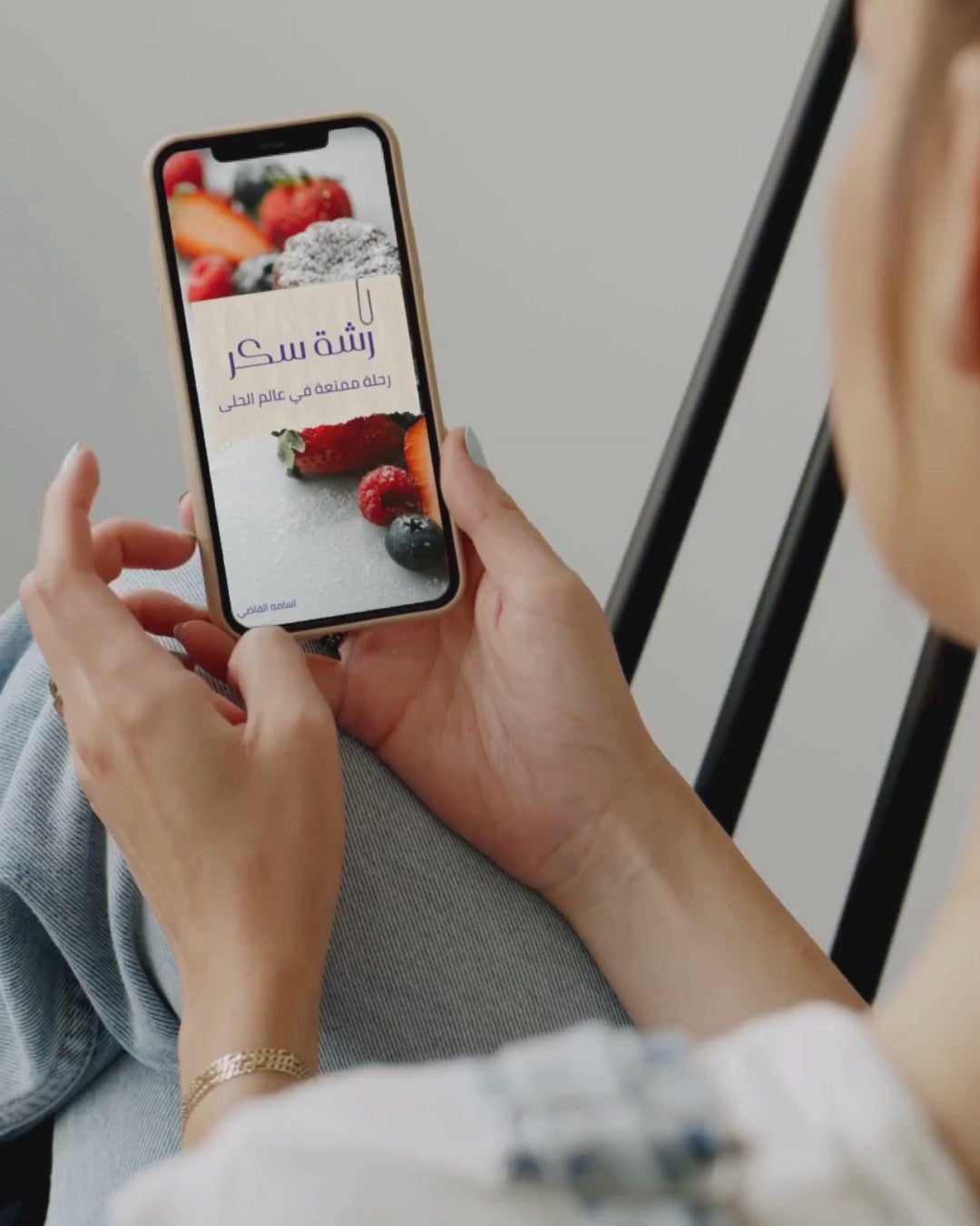 امرأة تمسك هاتفًا يعرض غلاف كتاب إلكتروني بعنوان 'رشة سكر | دليلك للحلويات الشرقية والغربية تعلم اجمل الوصفات' مع صورة لحلويات بالتوت والسكر البودرة.